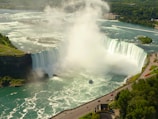 A large, powerful waterfall with cascading water surrounded by lush green landscapes. Mist rises from the bottom, where a tour boat sails close to the falls. The scene includes nearby cliffs and a winding road with people walking along it.