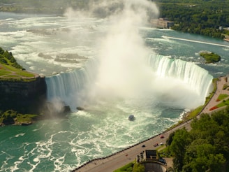 A large, powerful waterfall with cascading water surrounded by lush green landscapes. Mist rises from the bottom, where a tour boat sails close to the falls. The scene includes nearby cliffs and a winding road with people walking along it.