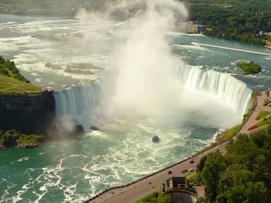 A large, powerful waterfall with cascading water surrounded by lush green landscapes. Mist rises from the bottom, where a tour boat sails close to the falls. The scene includes nearby cliffs and a winding road with people walking along it.