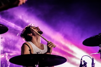 A drummer is energetically performing, with a backdrop of vibrant purple and pink stage lights creating a dynamic and intense atmosphere. The musician is in a sleeveless top, passionately immersed in the music.
