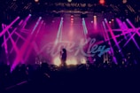 A vibrant concert scene with bright, multicolored stage lights illuminating a performer holding a microphone. The backdrop displays large, stylized text. The audience is silhouetted in the foreground, creating a dynamic atmosphere.