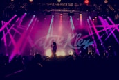 A vibrant concert scene with bright, multicolored stage lights illuminating a performer holding a microphone. The backdrop displays large, stylized text. The audience is silhouetted in the foreground, creating a dynamic atmosphere.