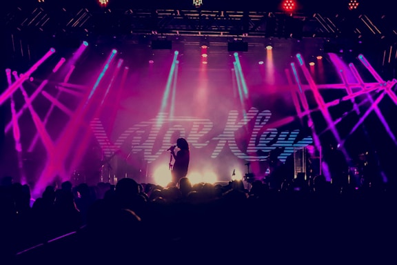 A vibrant concert scene with bright, multicolored stage lights illuminating a performer holding a microphone. The backdrop displays large, stylized text. The audience is silhouetted in the foreground, creating a dynamic atmosphere.