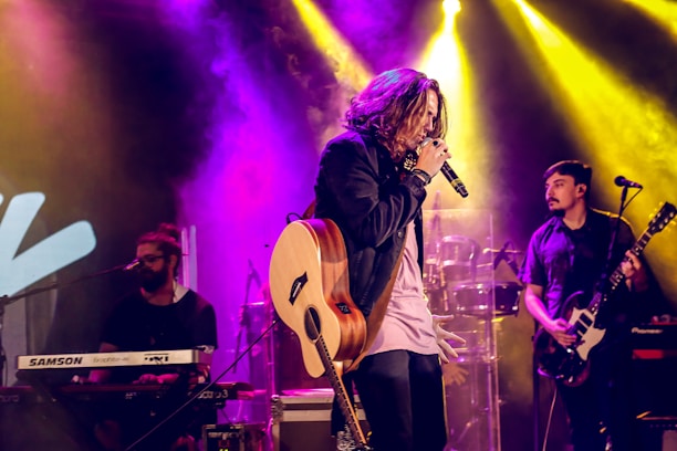 A musical performance is taking place on a stage with vibrant lighting. The central figure, holding a microphone and guitar, is energetically singing. There are musicians in the background playing keyboard and electric guitar. Colorful stage lights in shades of yellow, purple, and blue illuminate the scene, creating an exciting atmosphere.