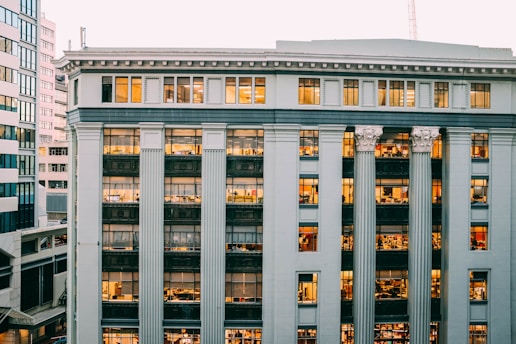 A multi-story office building with classical architectural elements such as large columns and detailed molding. The windows are lit from within, revealing numerous office spaces bustling with activity. The building's facade is a pale gray color, and it is surrounded by other urban structures.