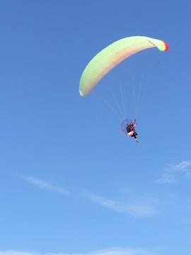 A person is paragliding against a clear blue sky. The paraglider wing is predominantly light green with a small section of red. The sky is mostly free of clouds, contributing to an open and airy atmosphere.