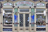 An ornate storefront with colorful tiles and decorative art, featuring the words 'A Perola do Bolhao'. Shelves are filled with various bottles, packages, and goods visible through the large glass windows. The facade includes detailed floral designs and illustrations of women in traditional attire.
