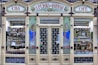An ornate storefront with colorful tiles and decorative art, featuring the words 'A Perola do Bolhao'. Shelves are filled with various bottles, packages, and goods visible through the large glass windows. The facade includes detailed floral designs and illustrations of women in traditional attire.