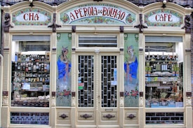 An ornate storefront with colorful tiles and decorative art, featuring the words 'A Perola do Bolhao'. Shelves are filled with various bottles, packages, and goods visible through the large glass windows. The facade includes detailed floral designs and illustrations of women in traditional attire.