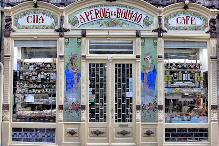 An ornate storefront with colorful tiles and decorative art, featuring the words 'A Perola do Bolhao'. Shelves are filled with various bottles, packages, and goods visible through the large glass windows. The facade includes detailed floral designs and illustrations of women in traditional attire.