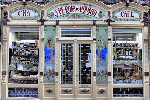 An ornate storefront with colorful tiles and decorative art, featuring the words 'A Perola do Bolhao'. Shelves are filled with various bottles, packages, and goods visible through the large glass windows. The facade includes detailed floral designs and illustrations of women in traditional attire.