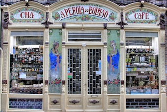 An ornate storefront with colorful tiles and decorative art, featuring the words 'A Perola do Bolhao'. Shelves are filled with various bottles, packages, and goods visible through the large glass windows. The facade includes detailed floral designs and illustrations of women in traditional attire.