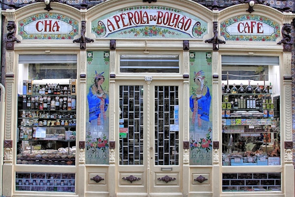 An ornate storefront with colorful tiles and decorative art, featuring the words 'A Perola do Bolhao'. Shelves are filled with various bottles, packages, and goods visible through the large glass windows. The facade includes detailed floral designs and illustrations of women in traditional attire.