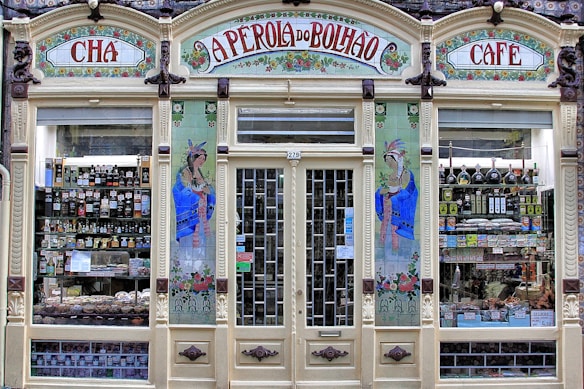 An ornate storefront with colorful tiles and decorative art, featuring the words 'A Perola do Bolhao'. Shelves are filled with various bottles, packages, and goods visible through the large glass windows. The facade includes detailed floral designs and illustrations of women in traditional attire.
