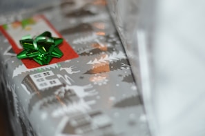 A wrapped gift featuring festive holiday-themed paper with white illustrations of reindeer, trees, and houses on a gray background. A shiny green bow is attached, along with a red tag.