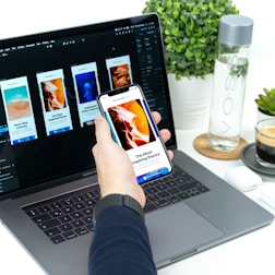A hand holding a smartphone displaying an article about inspiring places, set in front of a laptop showing a design application with multiple mobile app screens. A water bottle, a small plant, and a cup of coffee are placed on the white desk.