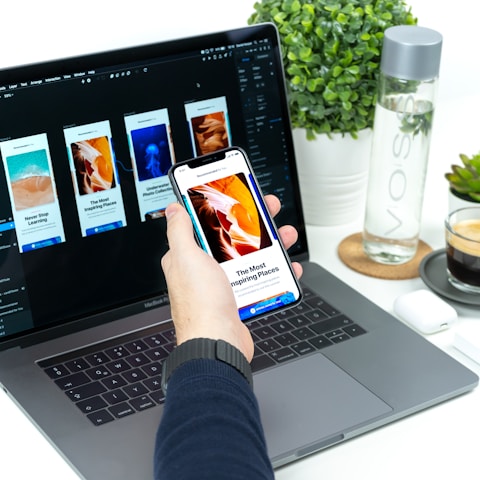 A hand holding a smartphone displaying an article about inspiring places, set in front of a laptop showing a design application with multiple mobile app screens. A water bottle, a small plant, and a cup of coffee are placed on the white desk.