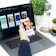 A hand holding a smartphone displaying an article about inspiring places, set in front of a laptop showing a design application with multiple mobile app screens. A water bottle, a small plant, and a cup of coffee are placed on the white desk.