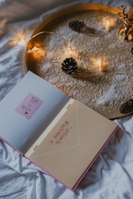 An open book with the words 'YOU are the BEST' on one page and 'A FAN LETTER TO MYSELF ...' on the other page is placed on a white cloth or bedspread. The background features a golden tray filled with white powder and decorated with small pinecones and a string of warm fairy lights.