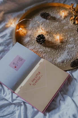 An open book with the words 'YOU are the BEST' on one page and 'A FAN LETTER TO MYSELF ...' on the other page is placed on a white cloth or bedspread. The background features a golden tray filled with white powder and decorated with small pinecones and a string of warm fairy lights.