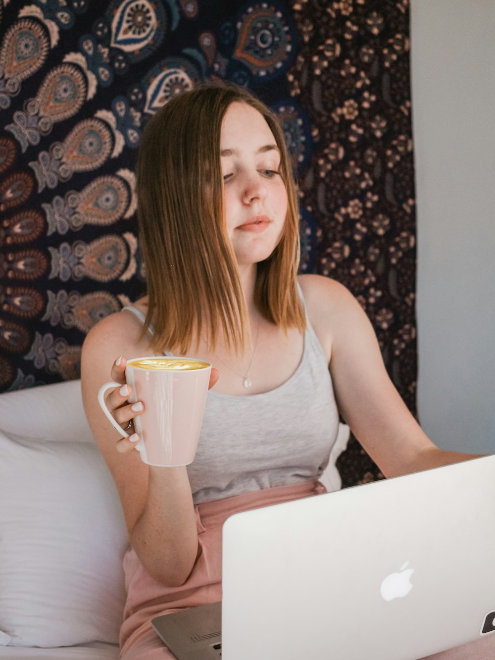 Frau benutzt MacBook und hält Tasse, während sie auf dem Bett sitzt