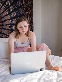 A young woman with light brown hair is sitting on a bed using a laptop. She appears focused and is casually dressed in a tank top and pink pants. The room has a tapestry with intricate patterns hanging on the wall behind her.