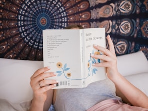 A person is lying on a bed reading a book titled 'the sun and her flowers' by Rupi Kaur. The book has a white cover with blue flowers. The background features a tapestry with intricate, circular patterns in shades of blue, orange, and white.