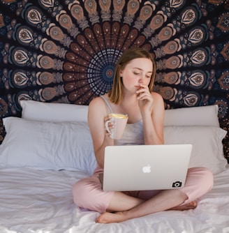 woman using MacBook and holding mug while sitting on bed