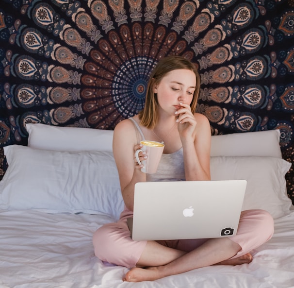 woman using MacBook and holding mug while sitting on bed