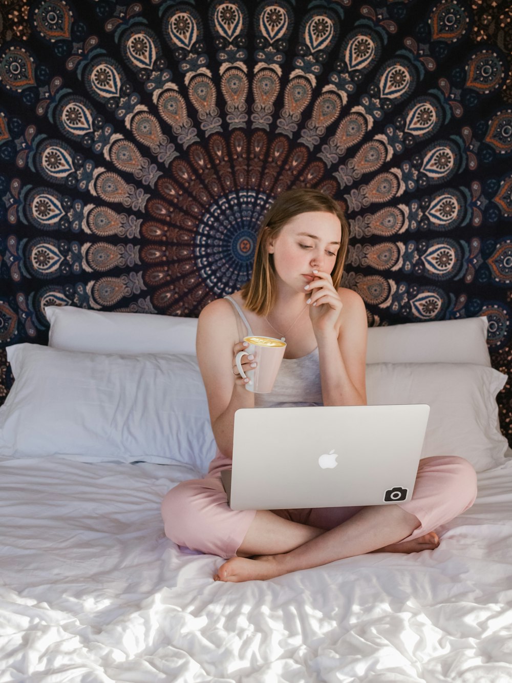 Frau benutzt MacBook und hält Tasse, während sie auf dem Bett sitzt