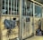 The image depicts the interior of a stable with wooden stalls. One stall has a horse inside, visible through the metal bars. Various equestrian equipment, including a saddle pad, bridle, and halter, are hanging on the wooden doors. The floor appears to be covered in sawdust or dirt, and a bag or backpack is placed on the ground on the right side.