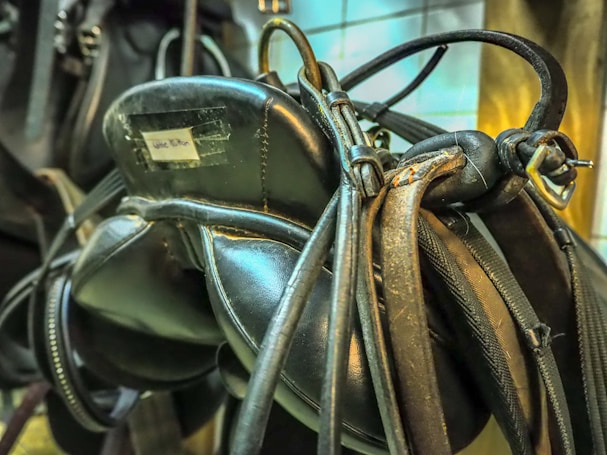 A close-up view of a well-worn leather saddle with straps and buckles, featuring a label that is slightly faded. The leather has a glossy, aged texture, indicating frequent use and care. The background shows a tiled wall with a warm light casting a glow over the scene.
