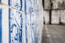 A close-up of a wall with decorative, blue and white tiles featuring intricate patterns. The tiles are aligned in rows alongside a blurred background of a roughly textured stone wall.