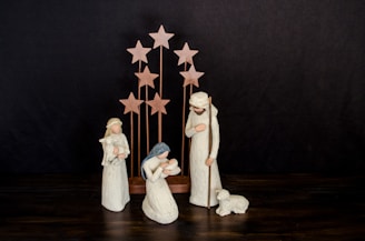 A nativity scene with three figurines representing traditional biblical figures. The figures are dressed in simple robes and are positioned around an infant. In the background, several wooden stars are arranged vertically, suggesting a celestial theme. A small lamb lies near one of the standing figures.