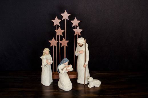 A nativity scene with three figurines representing traditional biblical figures. The figures are dressed in simple robes and are positioned around an infant. In the background, several wooden stars are arranged vertically, suggesting a celestial theme. A small lamb lies near one of the standing figures.