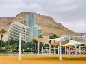A modern hotel building with a glass facade and a distinctive shape is set against the backdrop of rocky mountains. In the foreground, there are white sunshades supported by pillars over a sandy area with palm trees around. The sky appears overcast, adding a muted color to the overall scene.