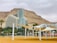 A modern hotel building with a glass facade and a distinctive shape is set against the backdrop of rocky mountains. In the foreground, there are white sunshades supported by pillars over a sandy area with palm trees around. The sky appears overcast, adding a muted color to the overall scene.