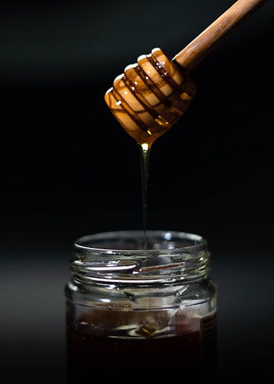 A close-up of golden honey dripping from a wooden dipper into a glass jar with wildflowers nearby.