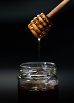 A honey dipper dripping golden honey into a glass jar, set against a dark, minimalistic background.