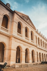 A grand, historical building with tall, arched windows and intricate architectural details. The facade is made of light-colored stone and features a sign reading 'Musée de l'Armée'. The sky above is blue with some clouds. Cobblestone pavement and cannons can be seen in the foreground.