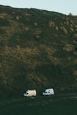 Two camper vans parked at the base of a large, grassy hill. The landscape is primarily natural, with shades of green dominating the scene. The terrain appears slightly rugged, with patches of grass and vegetation covering the hill.