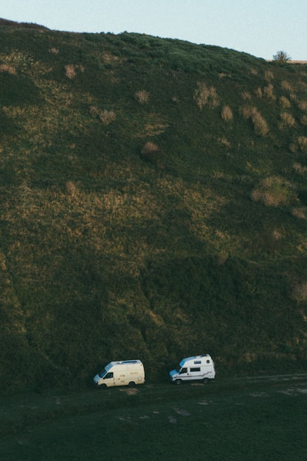 Two camper vans parked at the base of a large, grassy hill. The landscape is primarily natural, with shades of green dominating the scene. The terrain appears slightly rugged, with patches of grass and vegetation covering the hill.