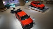 A sleek, modern showroom features a bright orange-red pickup truck and an SUV on display platforms. To the back left, a vintage turquoise truck is parked nearby a seating arrangement with a round table and several black chairs. The environment is spacious and has a polished, futuristic aesthetic with digital screens and elegant lighting.