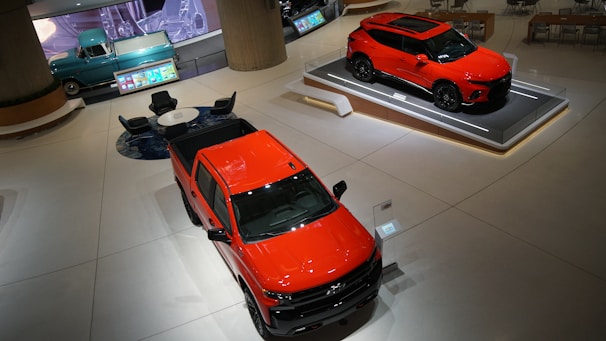 A sleek, modern showroom features a bright orange-red pickup truck and an SUV on display platforms. To the back left, a vintage turquoise truck is parked nearby a seating arrangement with a round table and several black chairs. The environment is spacious and has a polished, futuristic aesthetic with digital screens and elegant lighting.