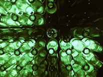 A collection of green glass bottles arranged in a symmetrical pattern, viewed from the top. The bottles are illuminated, casting a glowing green light. The setup creates a visually intriguing, repetitive pattern, with the circular openings of the bottles prominently featured.