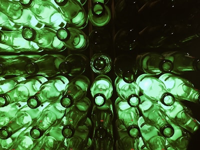 A collection of green glass bottles arranged in a symmetrical pattern, viewed from the top. The bottles are illuminated, casting a glowing green light. The setup creates a visually intriguing, repetitive pattern, with the circular openings of the bottles prominently featured.