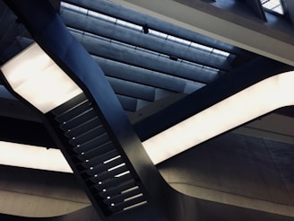 Detailed view of a modern architectural interior featuring an intertwining set of black metal beams and rectangular light fixtures. Overhead, there is a ceiling with geometric cutouts and diffused natural light filtering through.