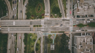 An aerial view of a busy city intersection with multiple roads converging at a central point. Vehicles are visible traveling in different directions, and there are sidewalks lined with buildings and green areas, which include manicured lawns and decorative landscaping.