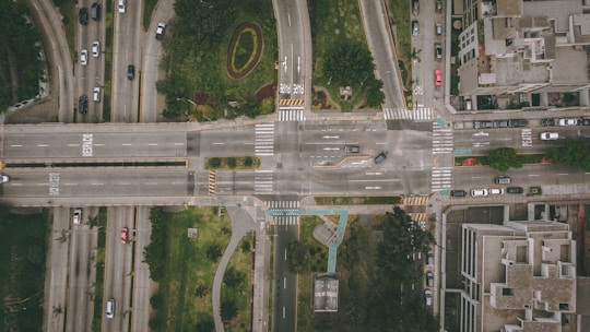An aerial view of a busy city intersection with multiple roads converging at a central point. Vehicles are visible traveling in different directions, and there are sidewalks lined with buildings and green areas, which include manicured lawns and decorative landscaping.