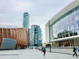 A modern urban setting featuring contemporary architectural designs. On the left is a distinctive building with curved, wooden paneling, suggesting an auditorium or cultural center. Adjacent high-rise buildings with balconies boast a sleek, residential look. The right side showcases a modern office building with large glass windows. Pedestrians walk along the spacious, clean concrete walkway under a cloudy sky.
